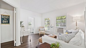 A living room with a white sofa, a wooden coffee table, and a beach-themed chair. There is a window with blinds and a lamp on a side table. - Charming 1-bed cottage in a Tropical Setting in West Palm Beach. Great location! (West Palm Beach)