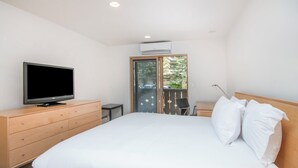 Una habitación de hotel moderna con una cama grande, un televisor en una cómoda de madera, una mesita y un balcón con vista a árboles.