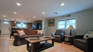 Amplia sala de estar con sofá, sillón y mesa de centro. Se ve una cocina al fondo, con armarios y un refrigerador.