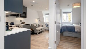 Een modern, compact appartement met kitchenette, een zithoek met een bank en fauteuil, en een slaapkamer met bed en raam.