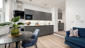 Een moderne keuken met grijze kastjes, een zwarte magnetron en een roestvrijstalen afzuigkap. Er is een zithoek met een zwarte tafel en blauwe stoelen, plus een blauwe bank met een kleurrijk kussen.