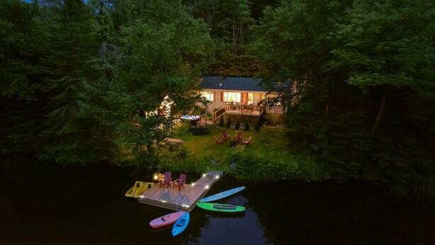 Una cabaña junto al lago rodeada de un bosque denso, con un muelle y kayaks.