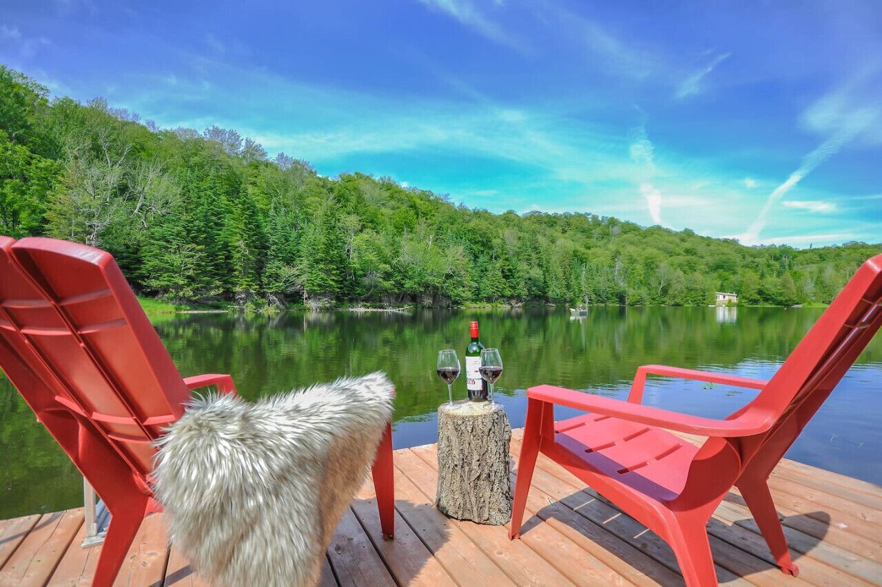 A wooden dock with red chairs and a bottle of wine on a tree stump.