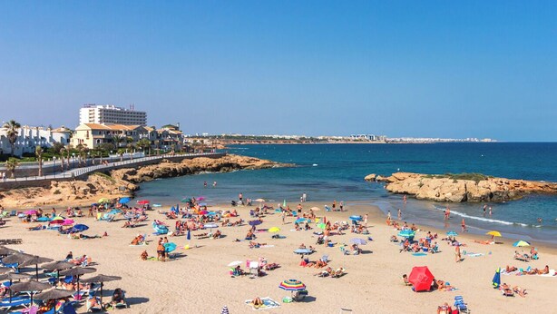 A beach with numerous umbrellas and people sunbathing. A hotel is visible in the background. - La casa Divertido (VC)