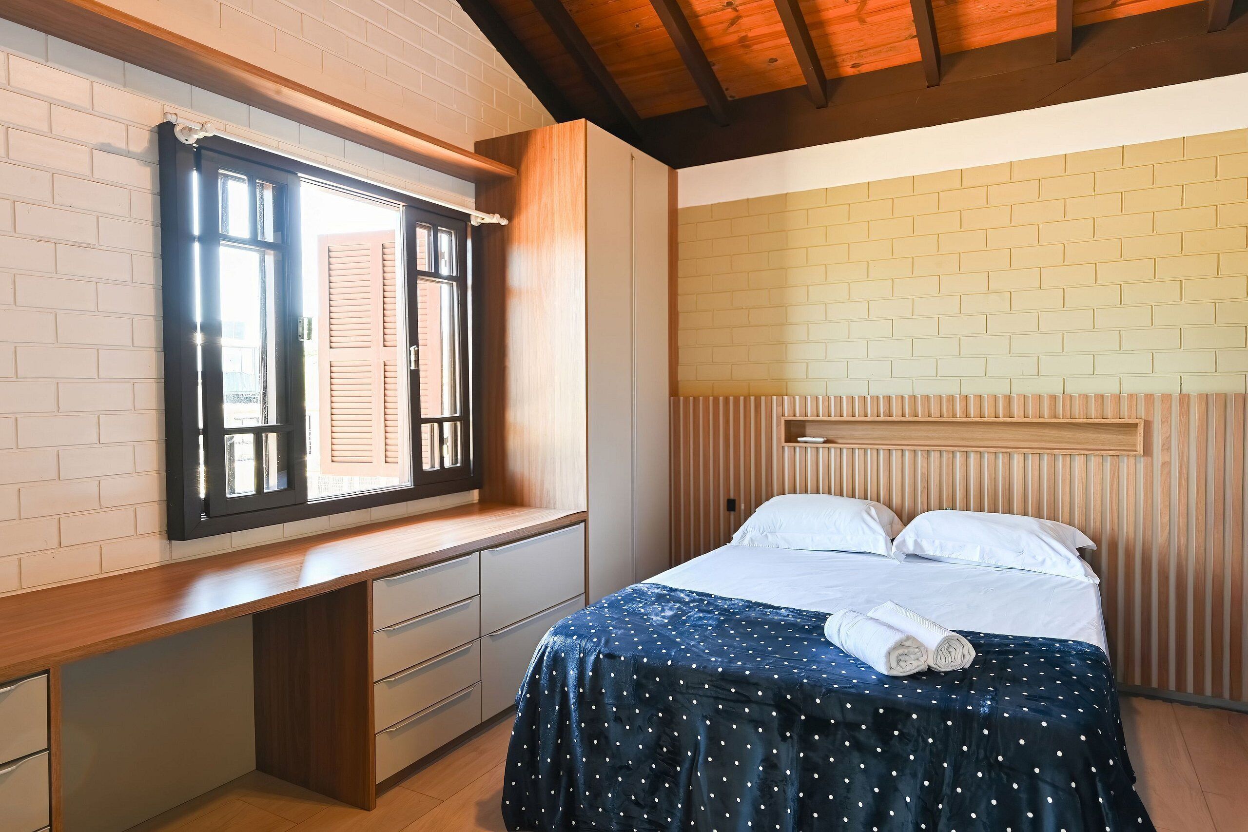 A bedroom with a wooden desk, a bed with a polka dot bedspread, and a window with shutters.