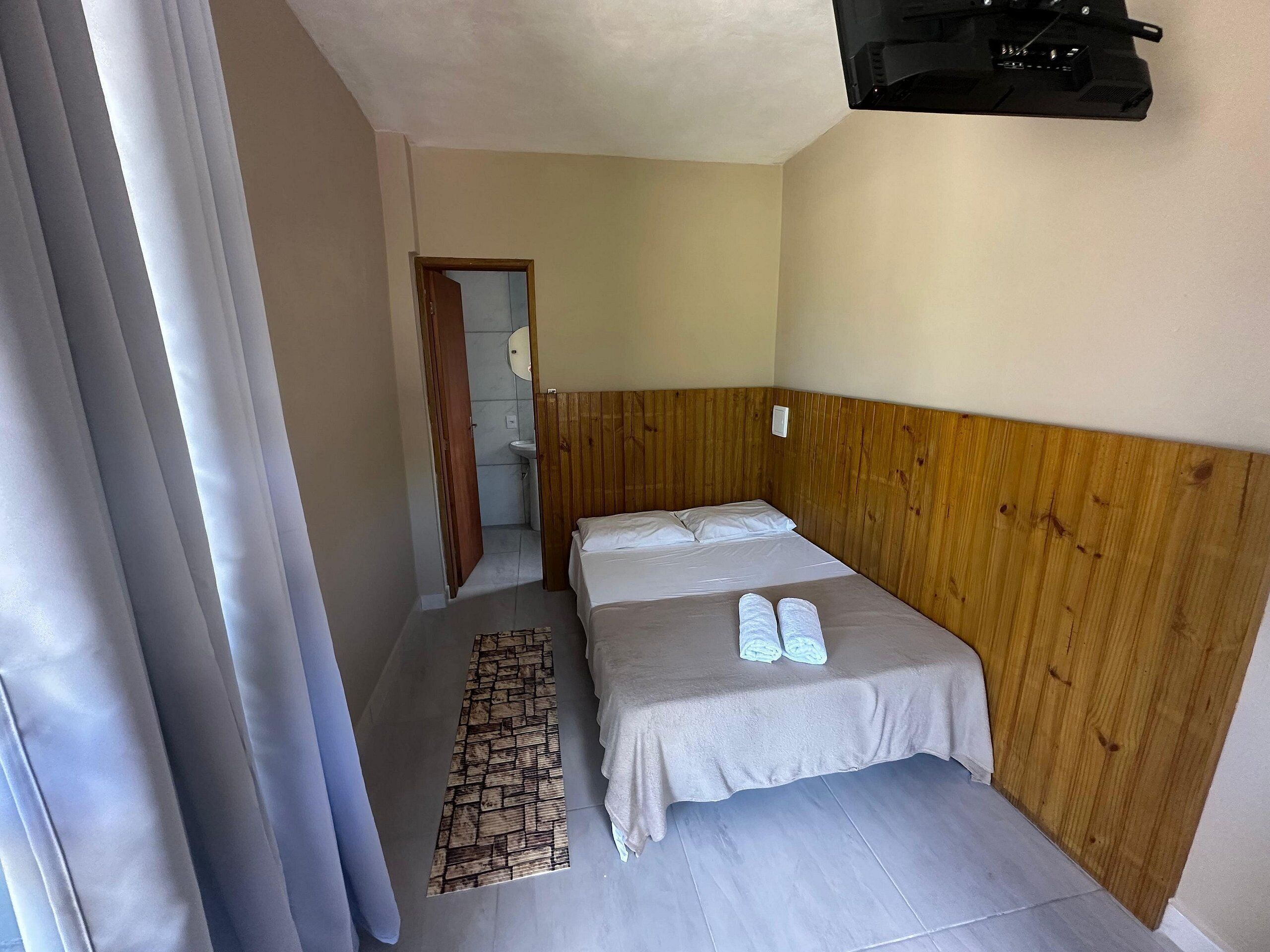 A small, neatly arranged room with a single bed, a wooden headboard, a small bedside table, and a wall-mounted TV.
