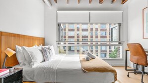 Une chambre d’hôtel moderne dotée d’une grande fenêtre, d’une tête de lit en bois, d’un lit avec du linge de maison blanc, d’un bureau avec une lampe et d’un fauteuil en cuir marron.