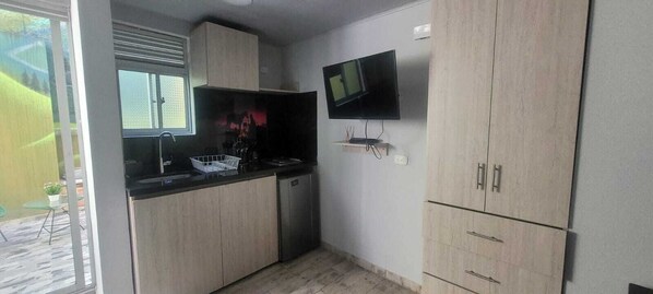 Une cuisine compacte équipée d’un lave-vaisselle intégré, d’un petit évier et d’un four à micro-ondes. On y trouve une fenêtre avec des stores, un téléviseur à écran plat fixé au mur, et un petit coin salon extérieur visible à travers la fenêtre.