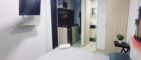 A compact bedroom with a bed, a small desk, a TV, and a kitchenette.