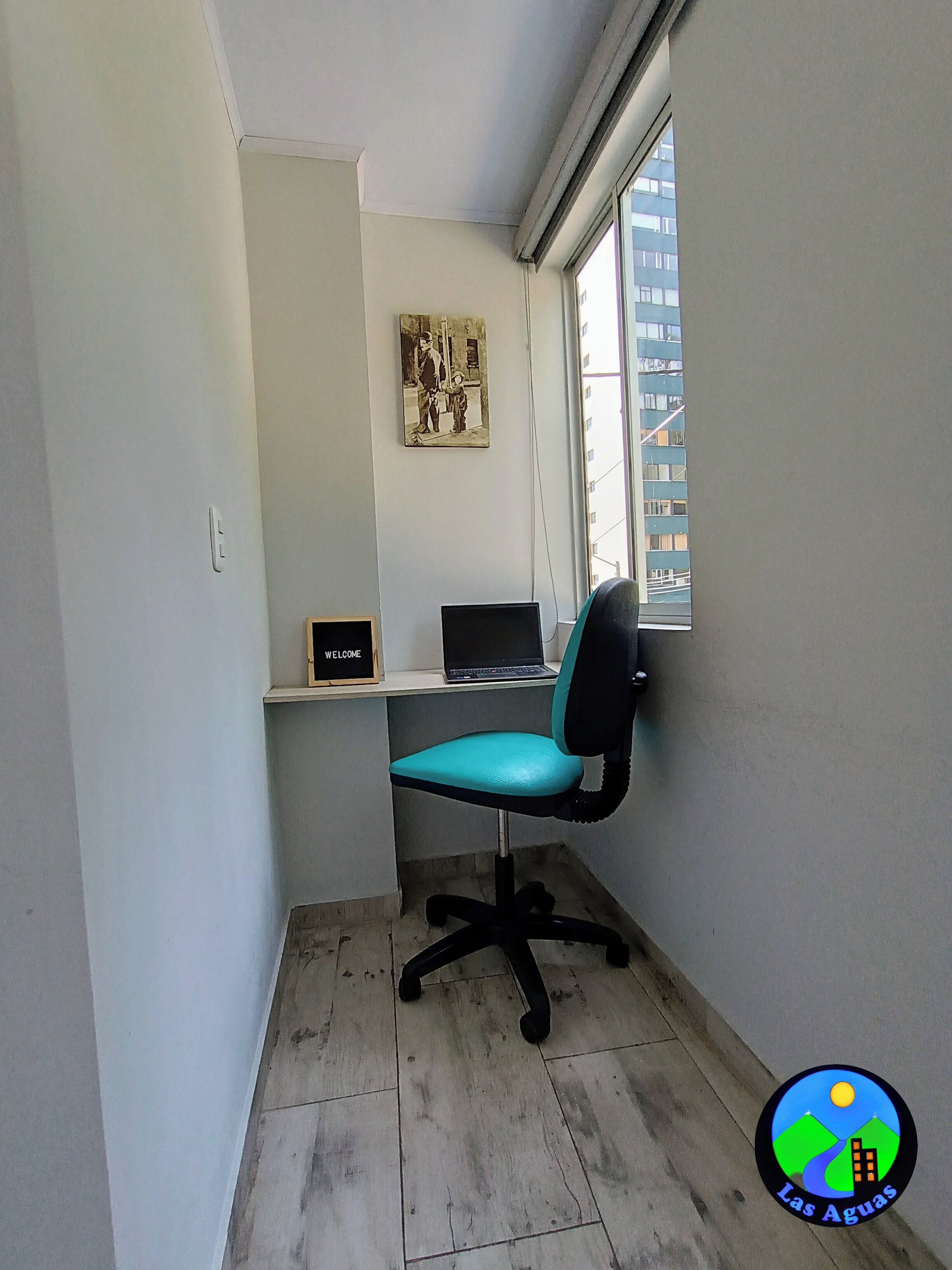 Un piccolo ufficio con una sedia da ufficio color petrolio, un portatile su uno scaffale e un quadro appeso al muro.