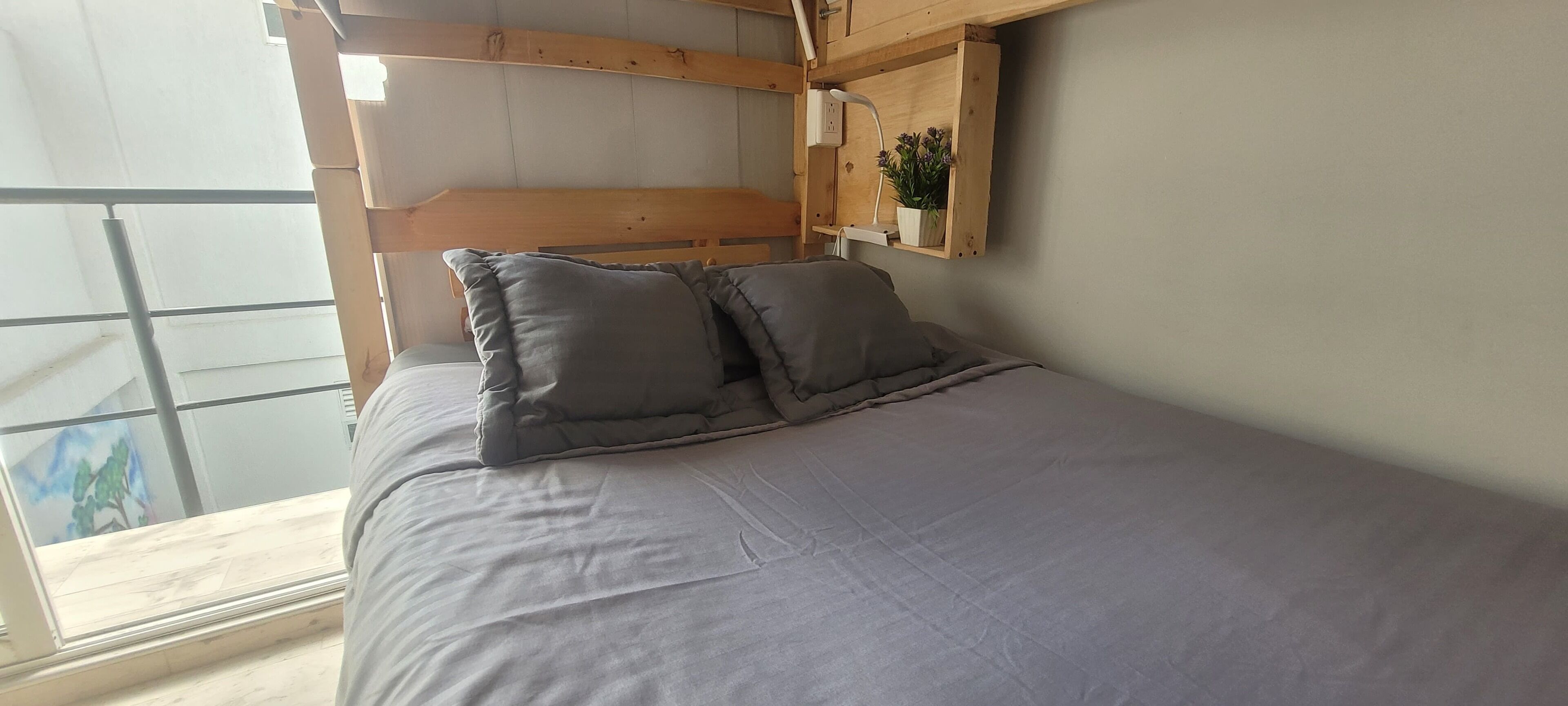 A bedroom with a bed, pillows, a wooden headboard, a shelf with a plant, and a window.