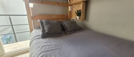 A bedroom with a bed, pillows, a wooden headboard, a shelf with a plant, and a window.