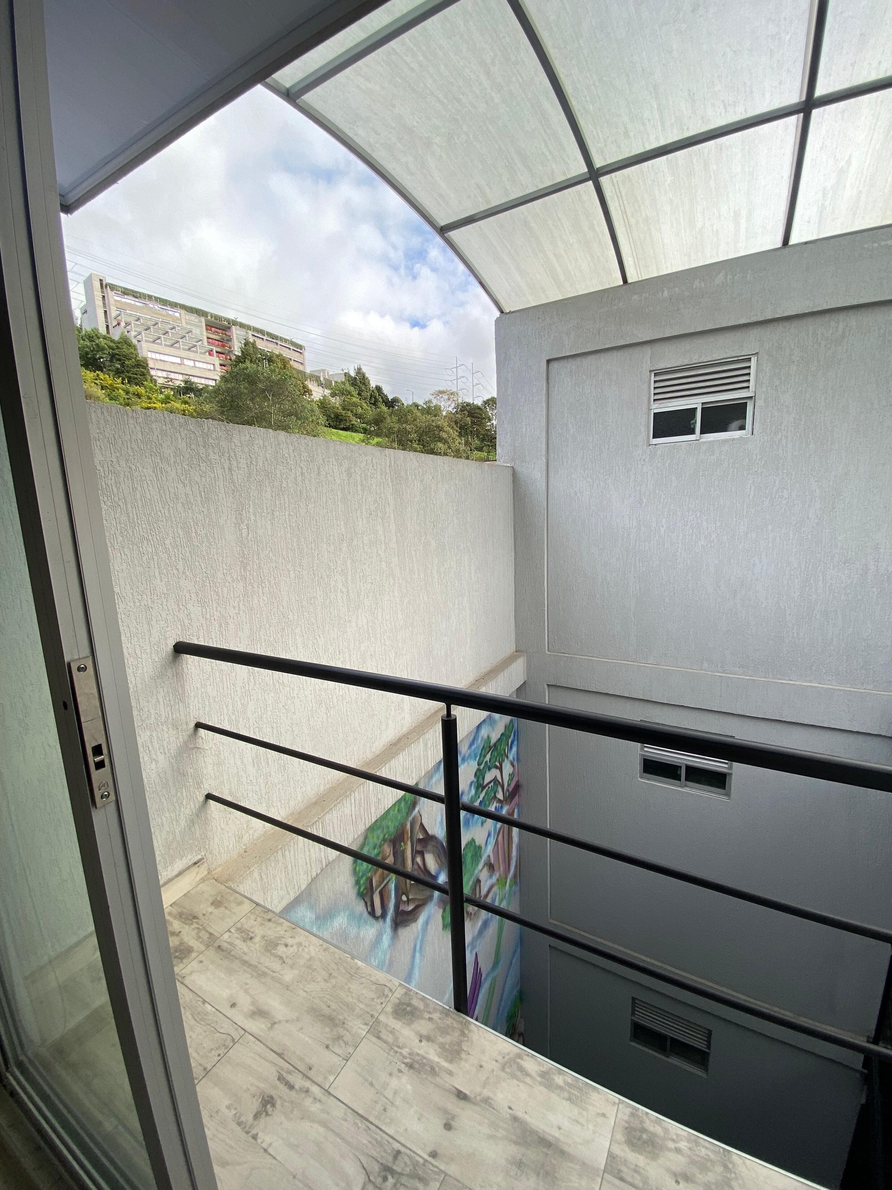 Un balcon doté d’une rambarde en métal, une vue sur le ciel et un bâtiment sur lequel est peinte une fresque en arrière-plan.