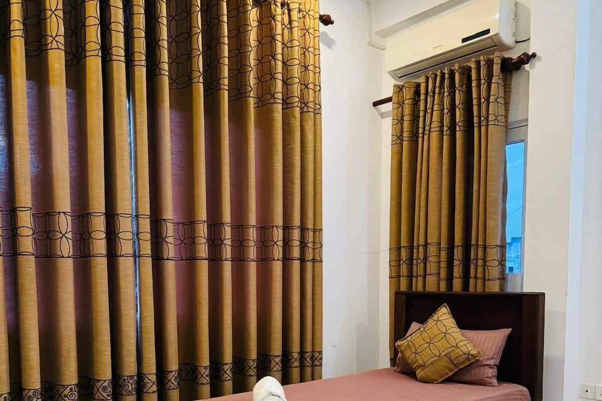 A single bed with a pink bedspread and a rolled towel, a wooden headboard, and a window with curtains.