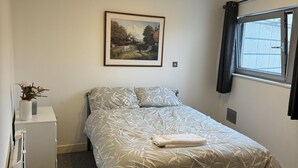A bedroom with a bed, a nightstand, a window with curtains, a painting on the wall, and a ceiling light fixture. - Bute Terrace Apartment - Cardiff City Center (Cardiff)