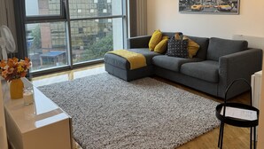 A modern living room with a grey sectional sofa, a black and white rug, a wooden floor, a white side table with a vase of flowers, and a large window with a cityscape view. - Bute Terrace Apartment - Cardiff City Center (Cardiff)