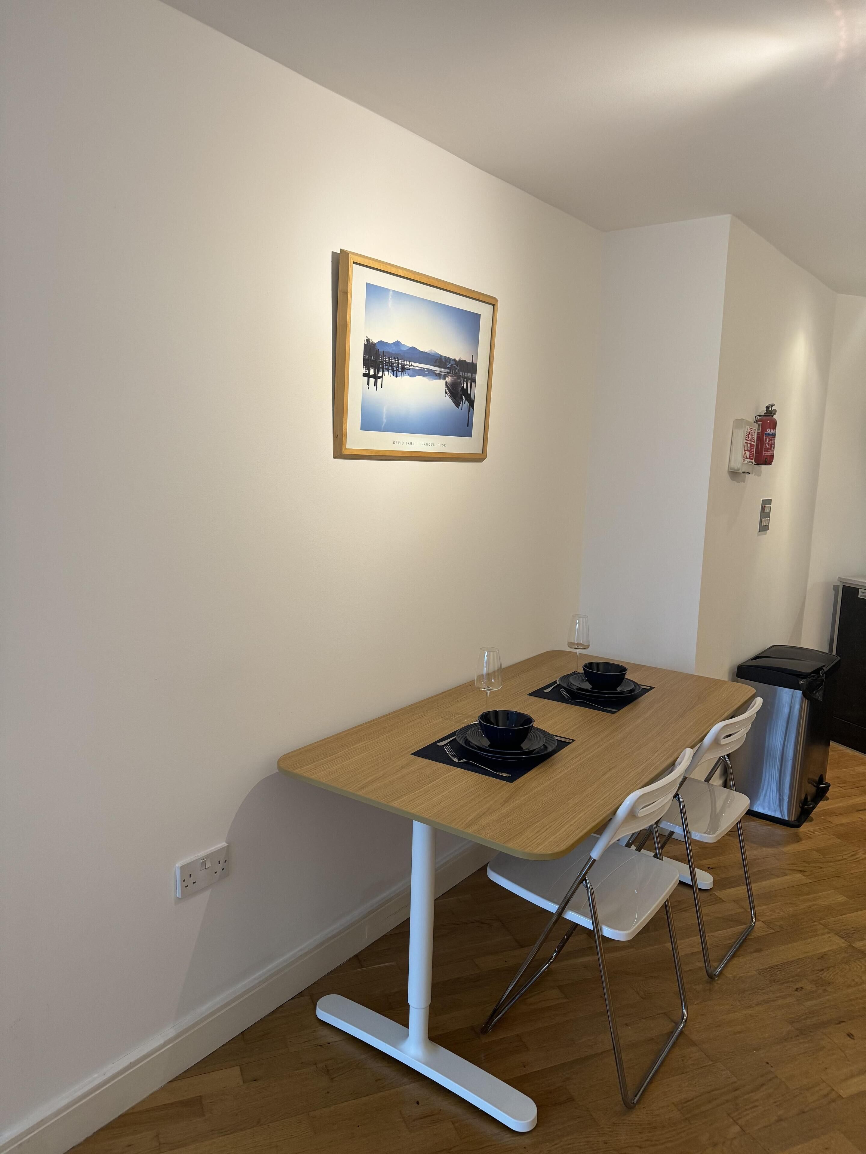 A dining area with a wooden table, white chairs, a framed photograph on the wall, and a fire extinguisher.