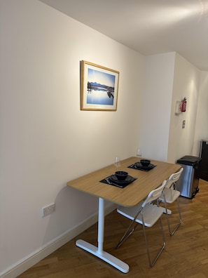A dining area with a wooden table, white chairs, a framed photograph on the wall, and a fire extinguisher.