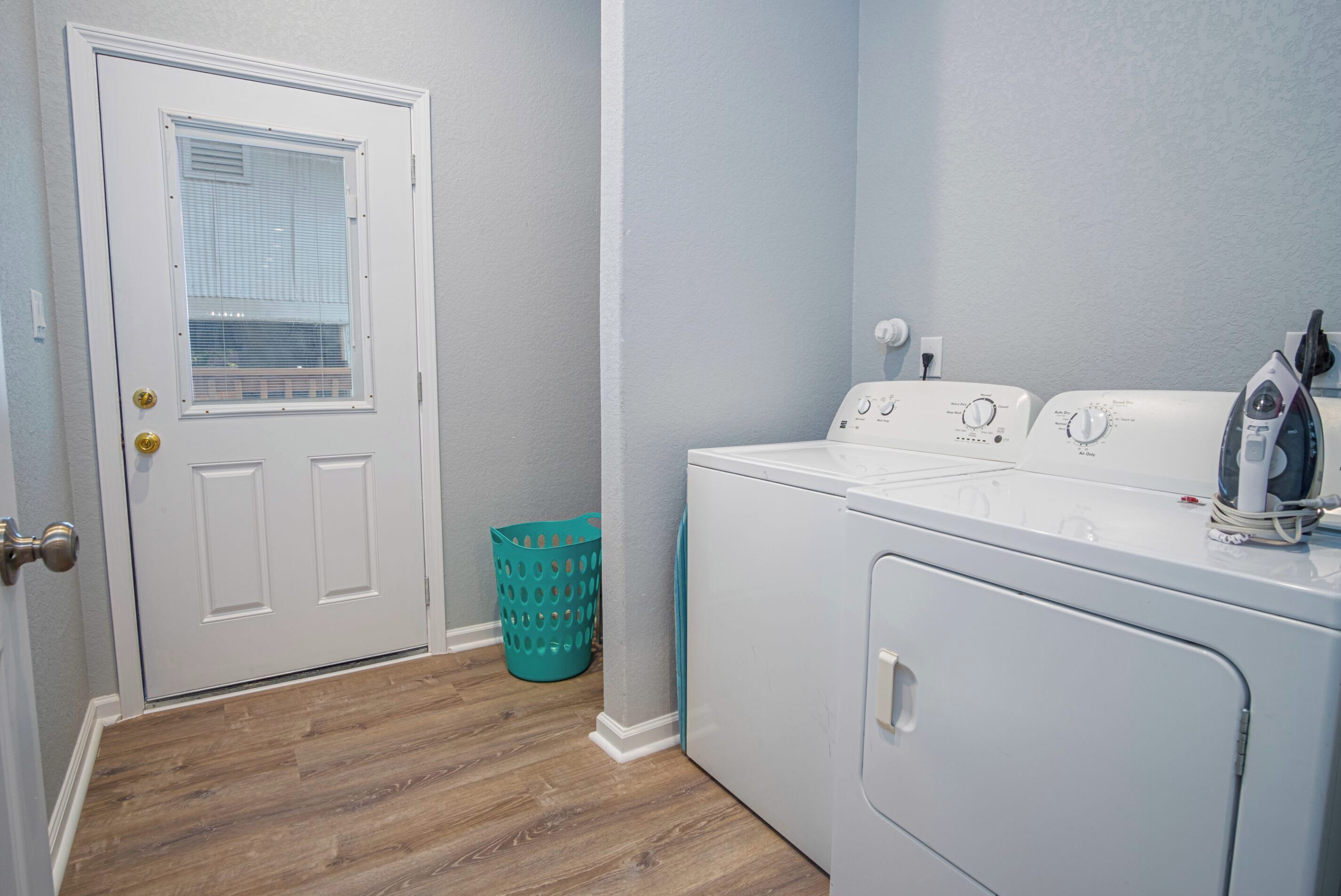 A laundry room with a front-loading washer and dryer, a door with a window, and a teal laundry basket.