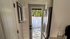Eine schmale Küche mit weißen Schränken, einem Edelstahl-Kühlschrank, einer Tür, die auf eine Terrasse führt, und einem kleinen Teppich auf dem Boden.