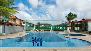 Uma piscina com um aviso 'Stay Tall', rodeada por palmeiras e edifícios com telhados de telha vermelha.