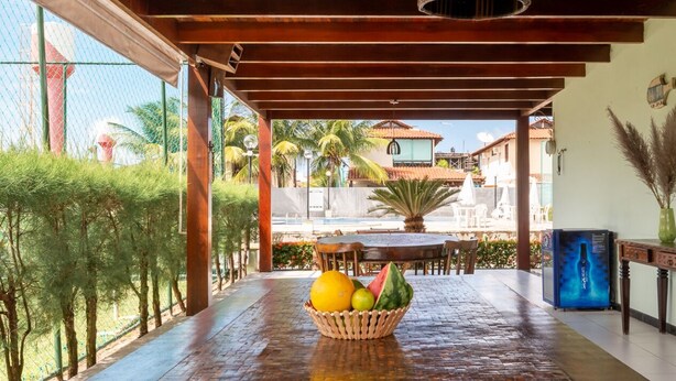 Uma área externa coberta com teto de madeira, uma mesa com cesta de frutas e vista para uma piscina e edifícios ao fundo.
