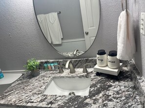 Ett badrum med en bänkskiva i granit, ett vitt handfat, en spegel och en handduksställning. På bänkskivan står två vita flaskor med etiketterna "Tvål" och "Lotion".