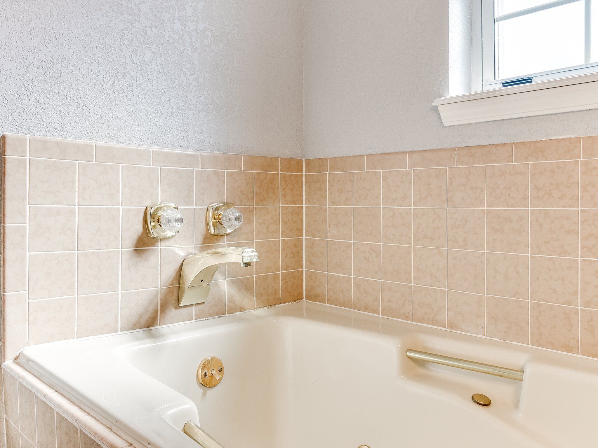 Et badeværelse med badekar, to haner og vægge beklædt med beige fliser.