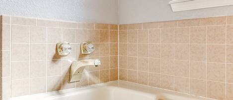 Et badeværelse med badekar, to haner og vægge beklædt med beige fliser.