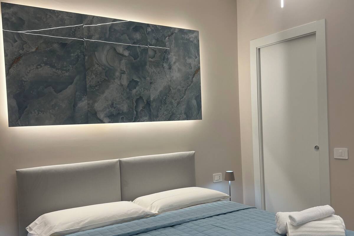 A modern bedroom with a large bed, a wall-mounted artwork, a bedside lamp, and a door.
