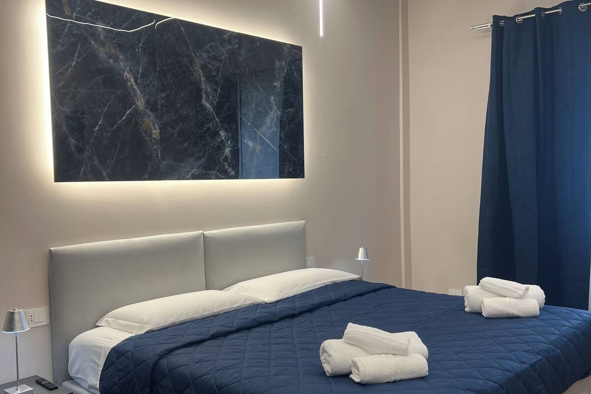 A modern hotel room with a large bed, a marble wall art piece, a nightstand with a lamp, and a blue curtain.