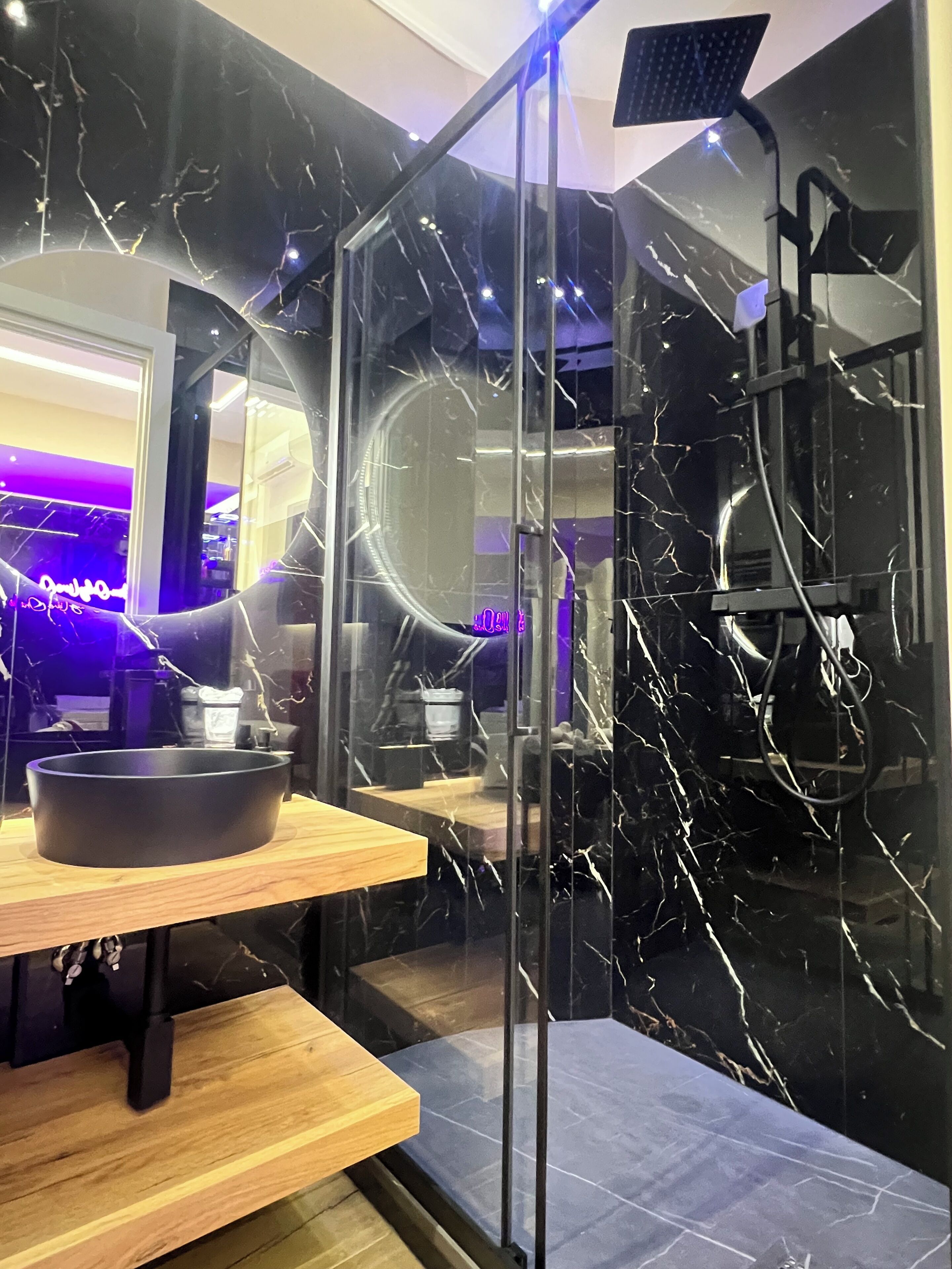 A modern bathroom with a black marble vanity, a wooden shelf, a round mirror, and a glass-enclosed shower area.