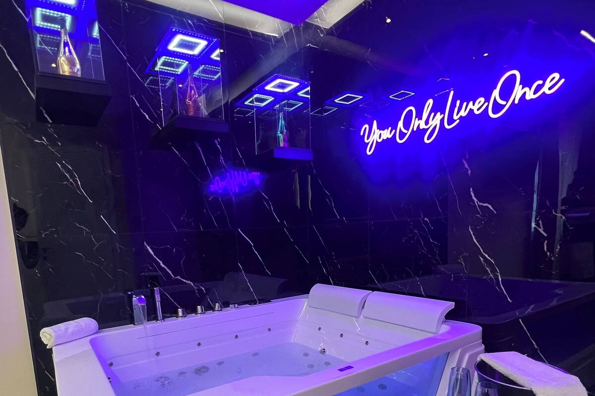 A modern spa area with a large white bathtub, a small round table with a glass, and a neon sign that reads "You Only Live Once".
