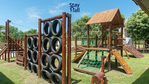 Un parque infantil con estructuras de neumáticos para escalar, una red de cuerdas y una casita de madera.