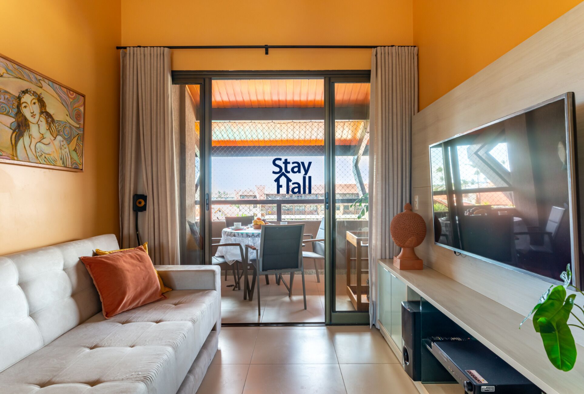 A modern hotel room with a sofa, a flat-screen TV, a dining table, and a sliding glass door leading to an outdoor area with a 'Stay Hall' sign.