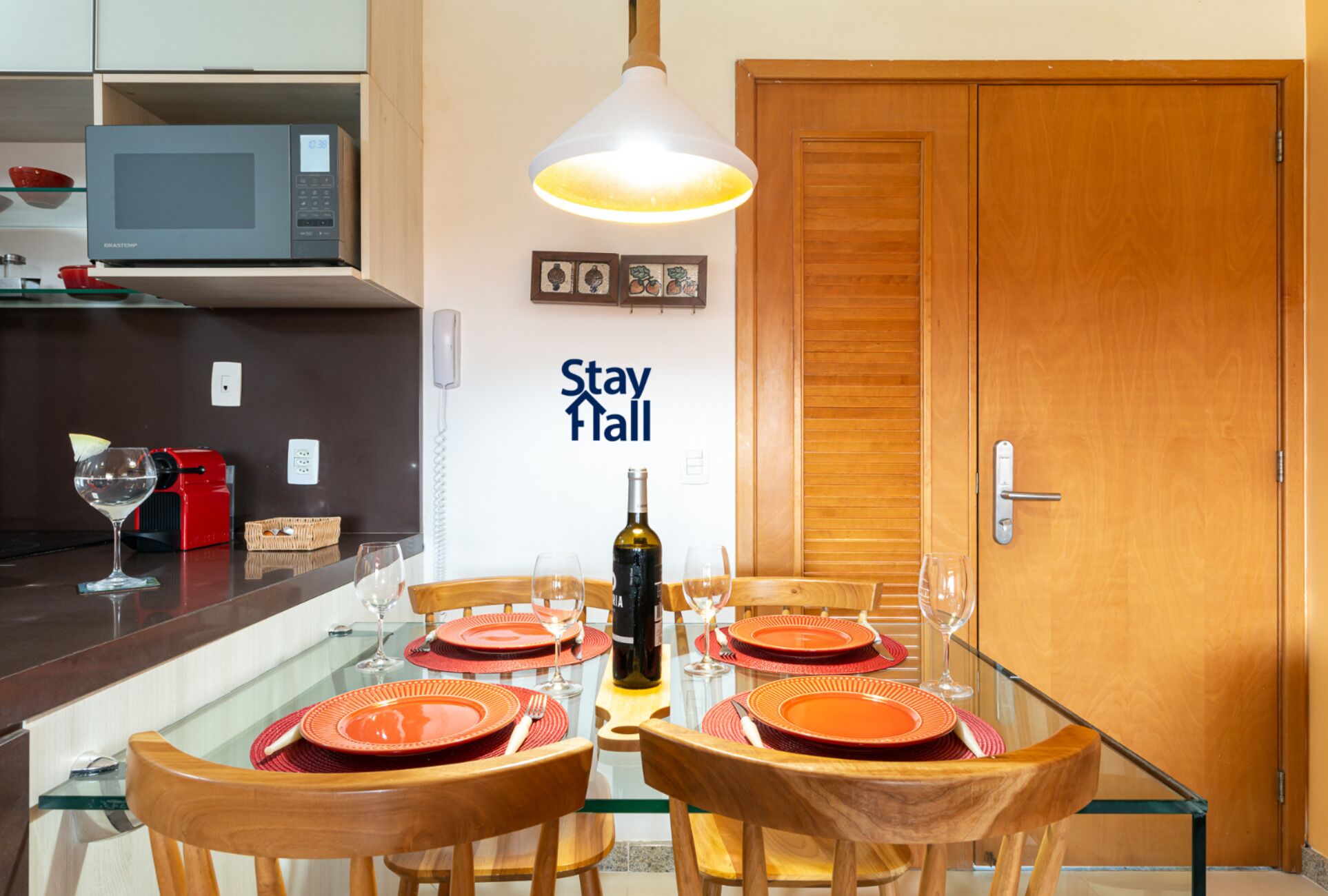 A dining area with a glass table, wooden chairs, and red plates. A bottle of wine and glasses are on the table. There is a microwave and a phone on the counter. A wooden door and a sign that reads "Stay Hall" are visible.