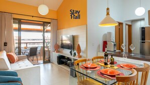 Una habitación de hotel moderna con una mesa de comedor de vidrio preparada para cuatro personas, un sofá, un televisor y una zona de cocina con un tostador y copas de vino.