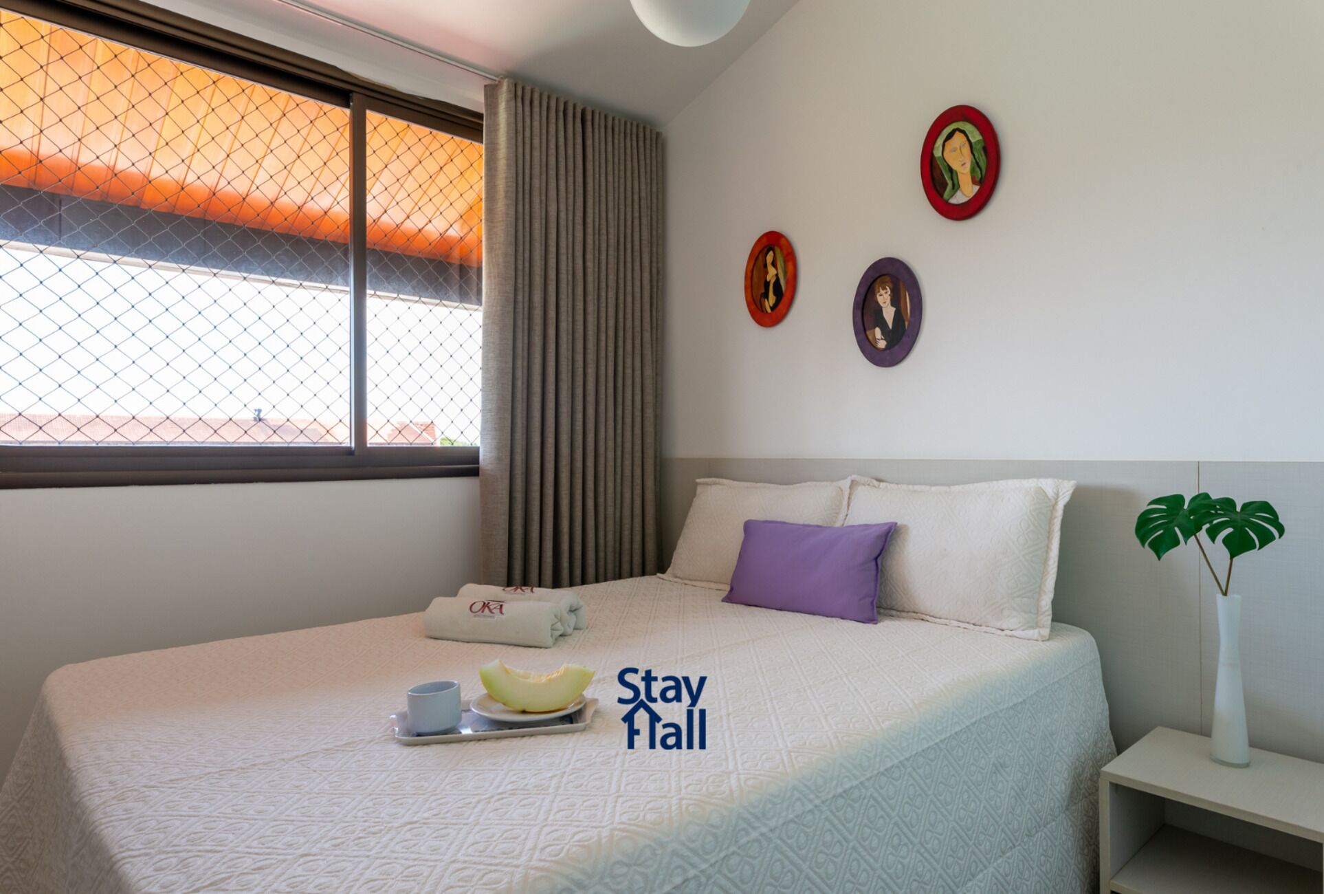 A hotel room with a bed, a tray with a cup and a fruit, a pillow, a plant, and artwork on the wall.