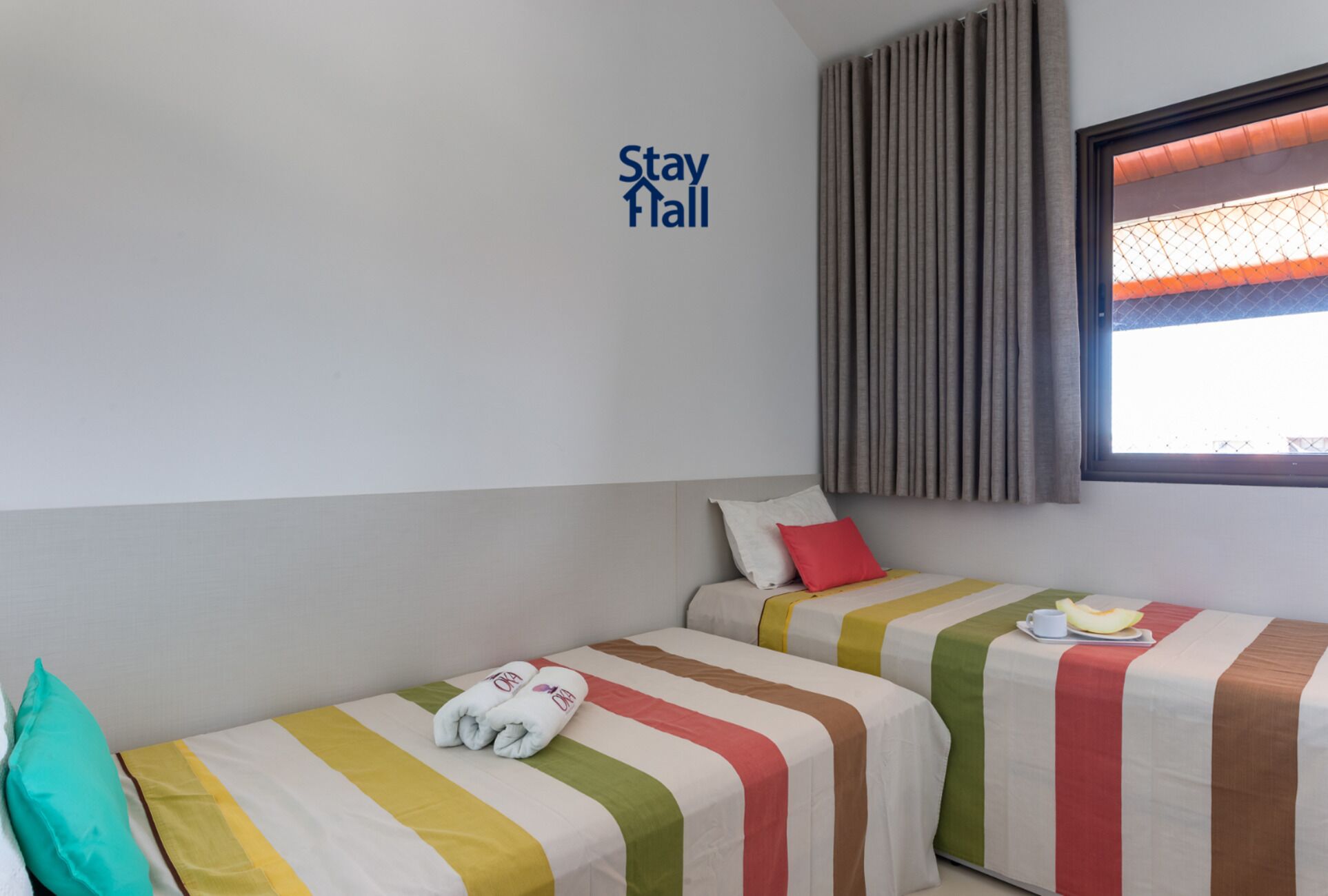 A hotel room with two beds, each with striped bedding and a pillow. A 'Stay Hall' sign is on the wall, and there is a window with curtains.