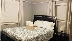 Una cama bien hecha con una colcha estampada, una mesita de noche con una lámpara y una cesta de toallas.