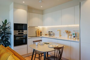 A modern kitchen with white cabinetry, a built-in oven, a dining table with chairs, and a potted plant. - Modern Studio Flat in North London (London)