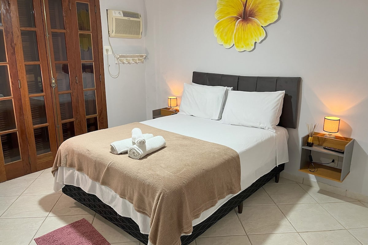 Un lit bien fait, avec une couverture marron, des oreillers blancs et une serviette enroulée posée sur le lit. On y voit également un climatiseur fixé au mur, une porte en bois avec des panneaux de verre et une décoration murale représentant une fleur jaune.