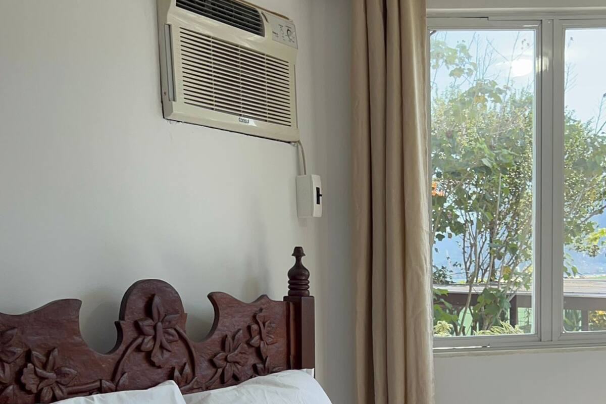 Un lit bien fait, avec du linge de lit blanc et un plaid marron, une tête de lit en bois ornée de motifs floraux, un climatiseur fixé au mur et une fenêtre avec des rideaux.
