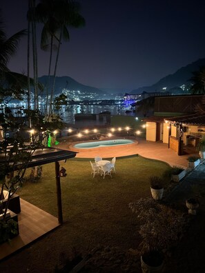 A nighttime view of a pool area with a dining setup and a view of a cityscape in the background. - Casa das Tartarugas (Angra dos Reis)