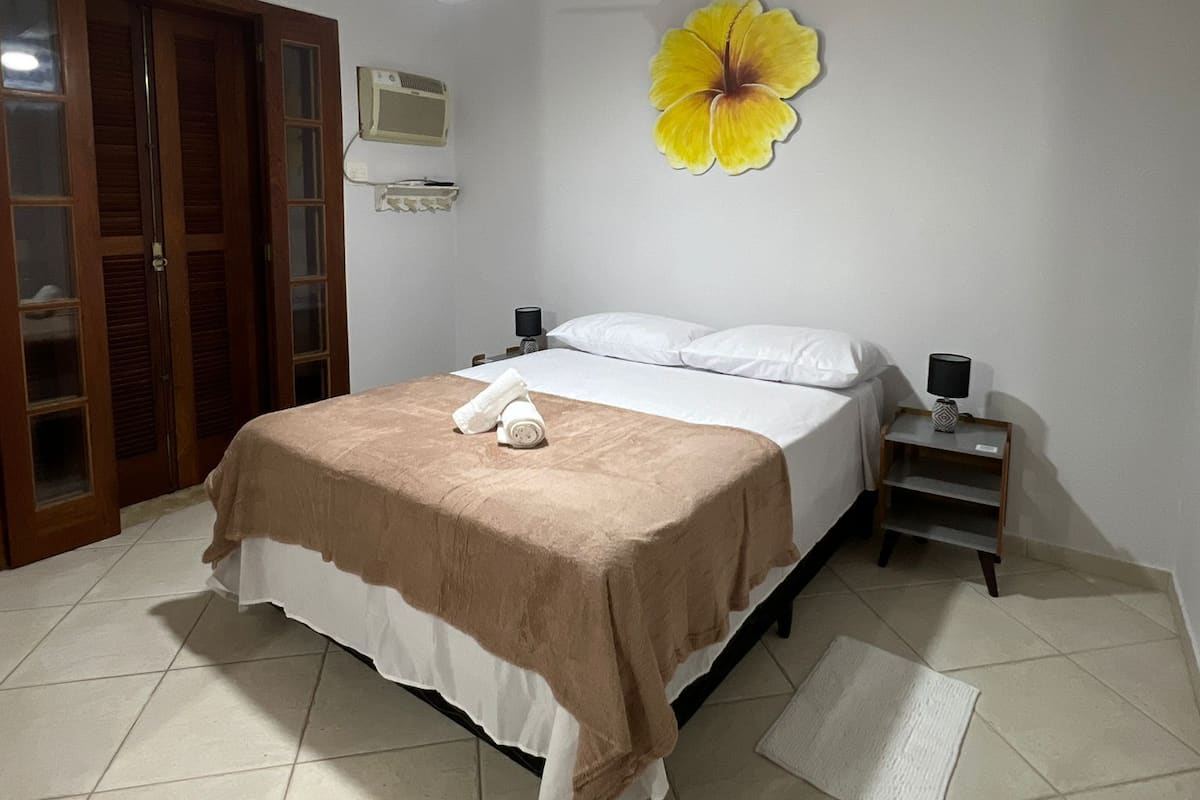 Une chambre à coucher comprenant un lit, une table de chevet, un ventilateur de plafond, un climatiseur mural et une porte en bois avec des panneaux de verre.