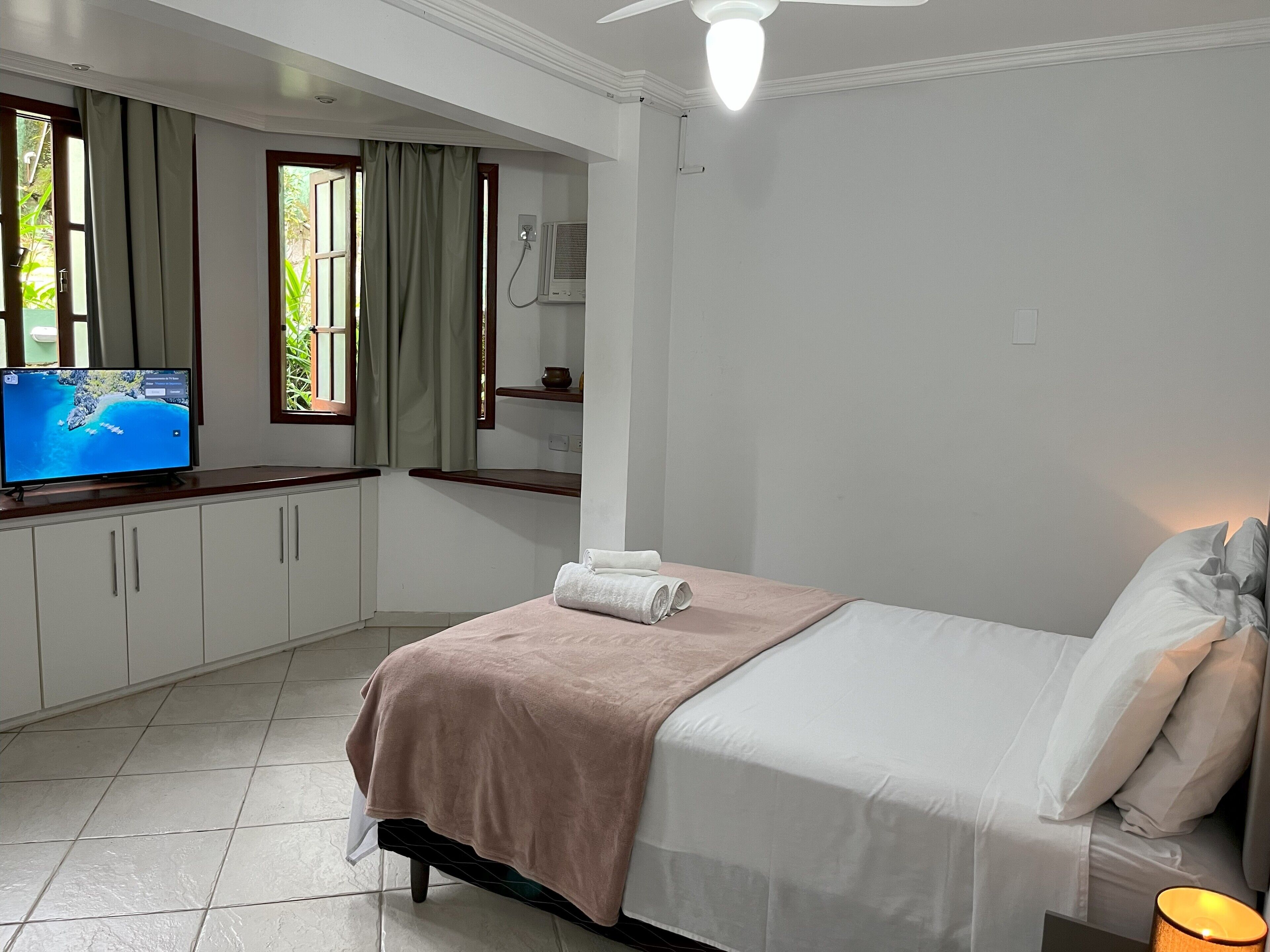 Een slaapkamer met een bed, een televisie op een bureau, een plafondventilator en een raam met gordijnen.