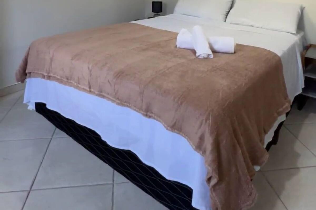 Un lit bien fait, recouvert d’une couverture marron et agrémenté d’oreillers blancs, une table de chevet avec une lampe et un ventilateur de plafond.