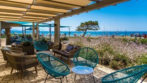Een terras buiten met rieten meubels en parasols, met uitzicht op een strand met boten en een heldere lucht.