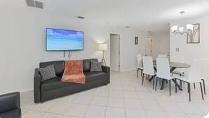 Ein modernes Wohnzimmer mit einer schwarzen Couch, einem an der Wand montierten Fernseher und einem Essbereich mit weißen Stühlen und einem Tisch.