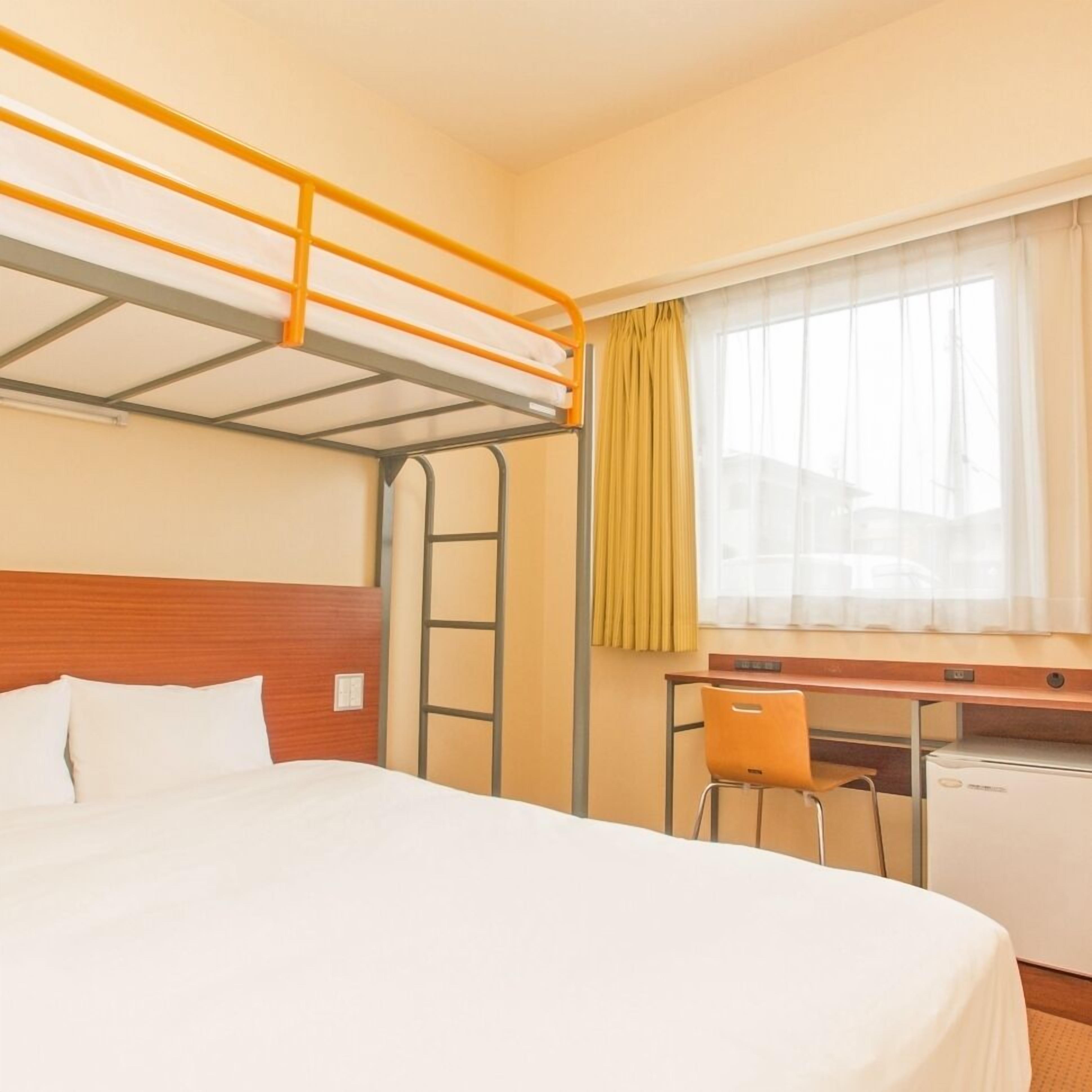 A hotel room with a bunk bed, a desk, a chair, and a small refrigerator.